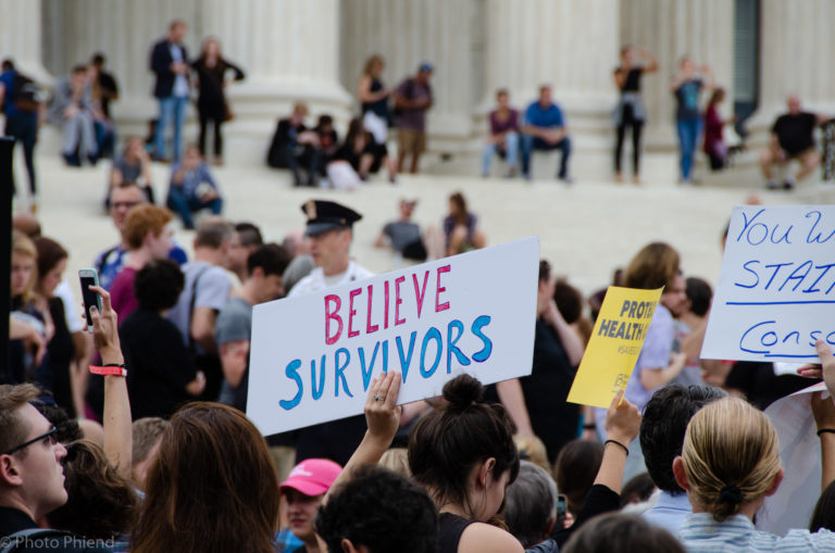 A crowd gathered outside with a woman holding up a sign that says Believe Survivors
